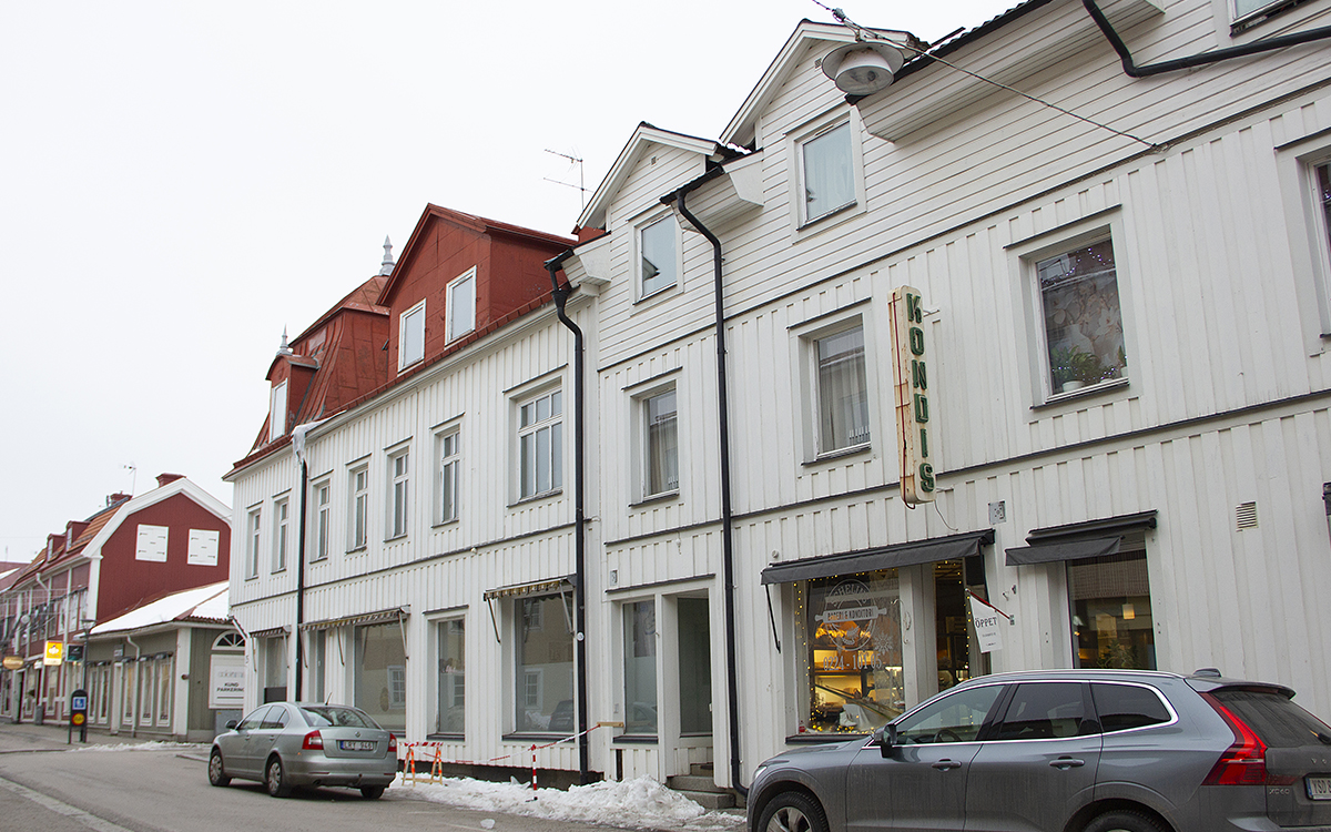 En gråvit huslänga med två hela våningar och en vindsvåning, en del av den i rött med röd takbeklädnad. Botten våningen har skyltfönster. Längst bort i bild är de frostade nedtill, närmast i bild används de för syltning och det finns en vit lite rostig släckt ljus-/neonskylt med grön vertikal text i versaler: KONDIS. På en liten flagga vid dörren intill står det "öppet". Fastigheten är en av dem som riskerar tvångsförsäljning på grund av AB-hems skulder hos Kronofogden.