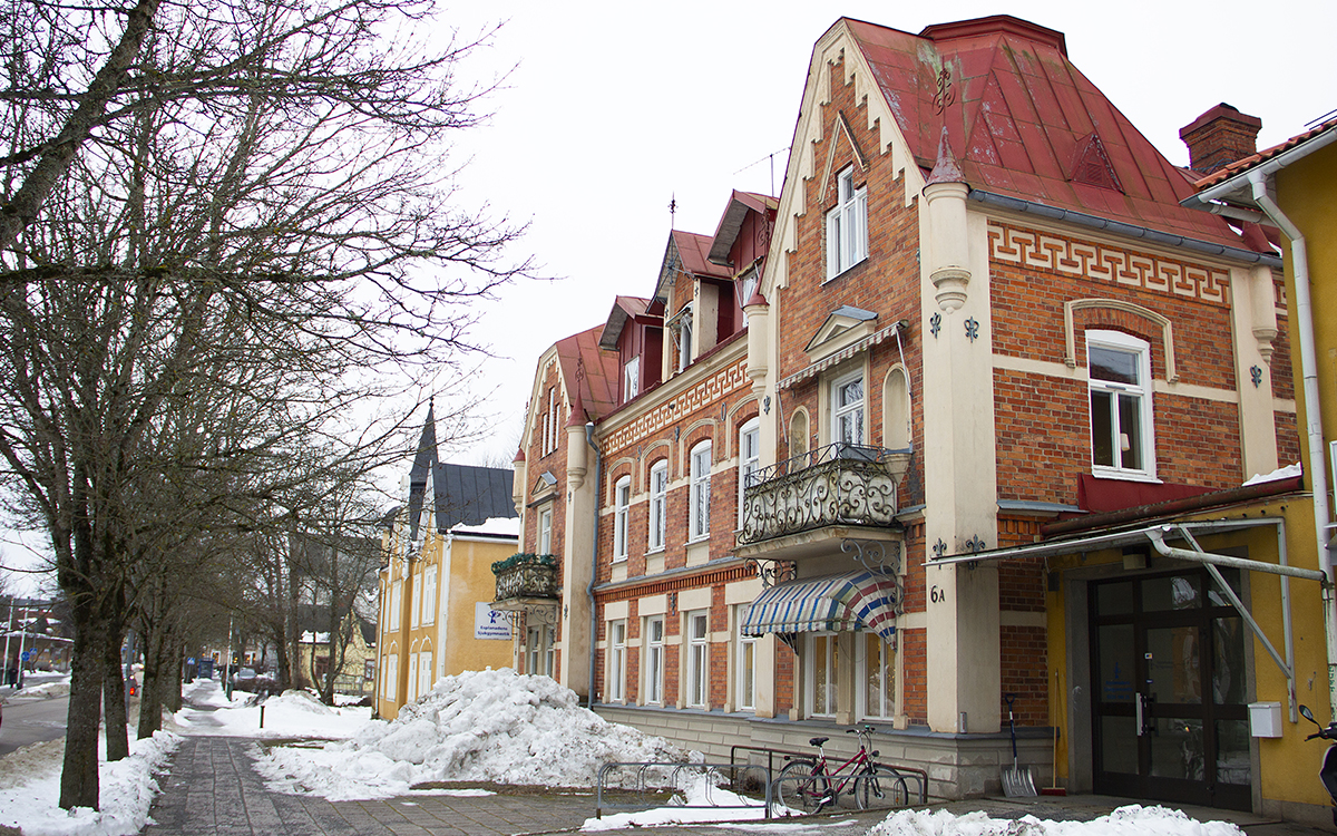 Sekelskifteshus i två våningar. En tegelvind dekorerad med lätta material, ett rött tak, ett litet dekorativt torn och två gjutjärnsbalkonger med invecklad dekoration. Framför huset låg ett berg av smutsig snö som tagits bort och en cykel parkerad. Trottoar och trädgräns. Detta hus på Nora Esplanad i Sala är ett av de bostäder som riskerar tvångsförsäljning på grund av skulder AB-home har hos Kronbödeln.