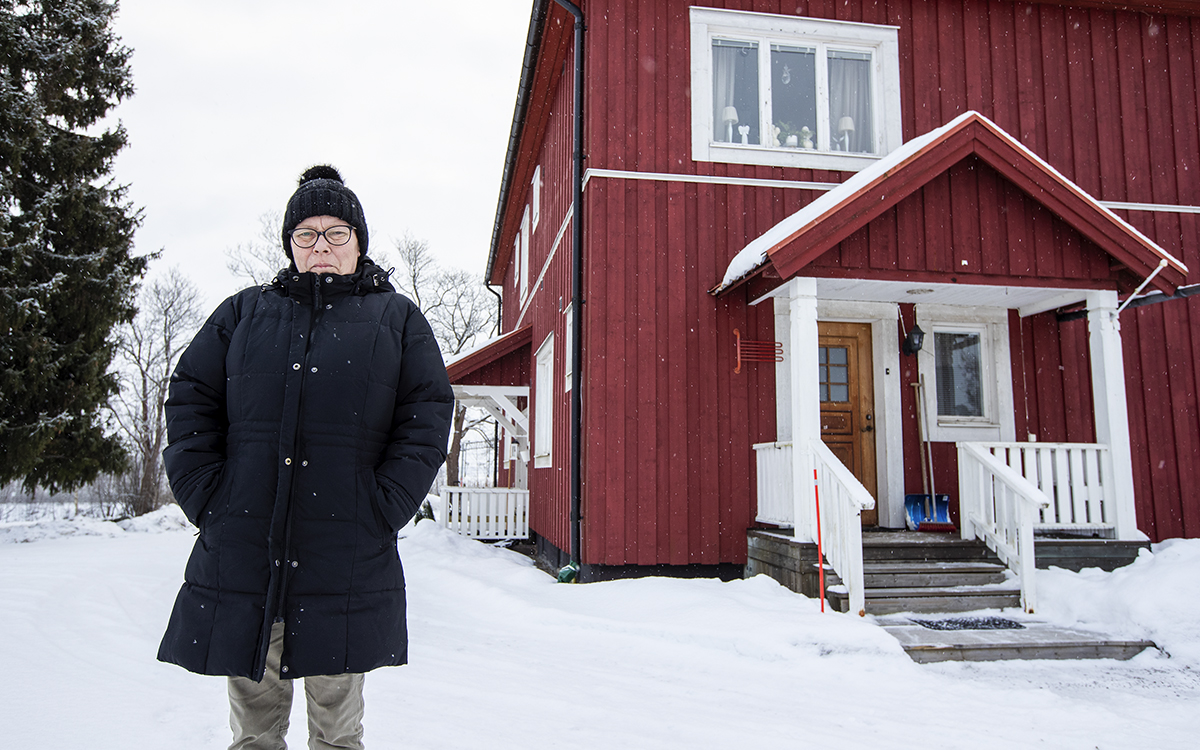 Sineve Raymondsdotter, till vänster, är klädd i svart/mörk vinterjacka och mössa, mörka glasögon och har händerna i fickorna. Bakom henne finns några snötäckta granar och kala träd. Till höger ligger ett tvåvånings rött trähus med vitt fönsterfoder och detaljer. Snö på marken, reflekterande stolpar för snöröjning i husets hörn. I en hyreslägenhet i AB-hem, Strvik, är det ett återkommande problem med att värmen går ut. Ägaren Annika Buckland skrev att trots de ekonomiska problemen går det mesta som vanligt när det kommer till att åtgärda felanmälningar.