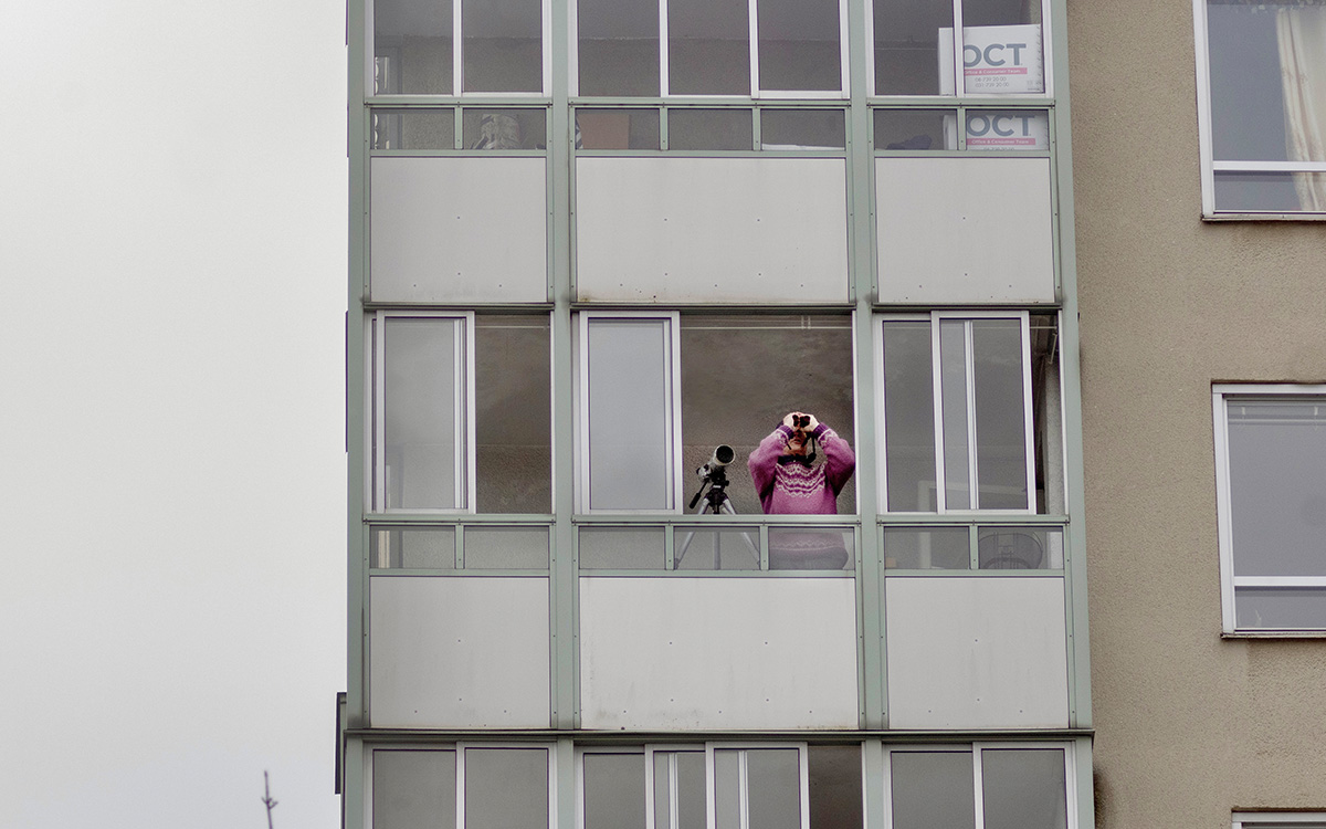 Fågelskådning från en balkong. Man med kikare och rosa tröja spanar mot himlen.