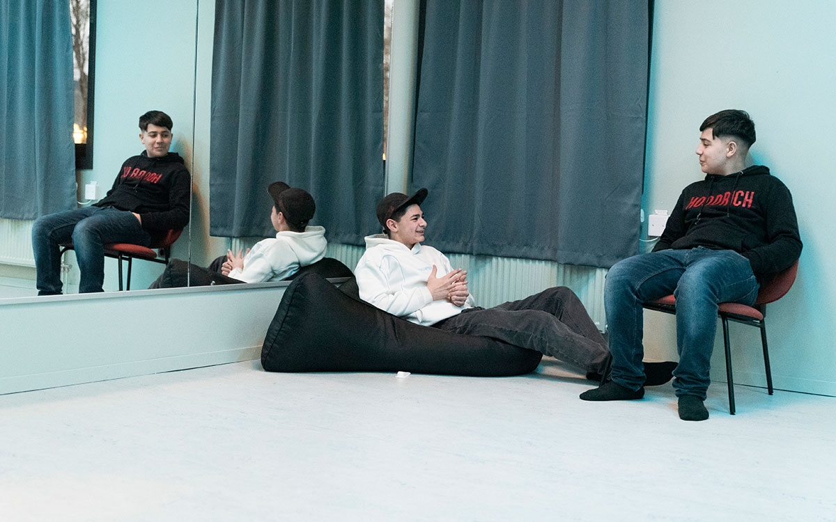 Två killar som sitter på en stol respektive sacco-säck i ett rum med blåaktigt ljus och en spegelvägg.