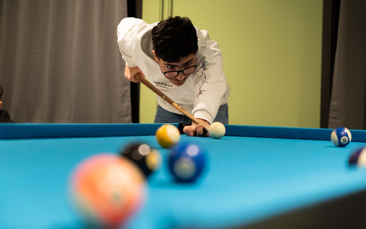 En ung kulle med kort mörkt hår, glasögon och vit tröja som spelar biljard.
