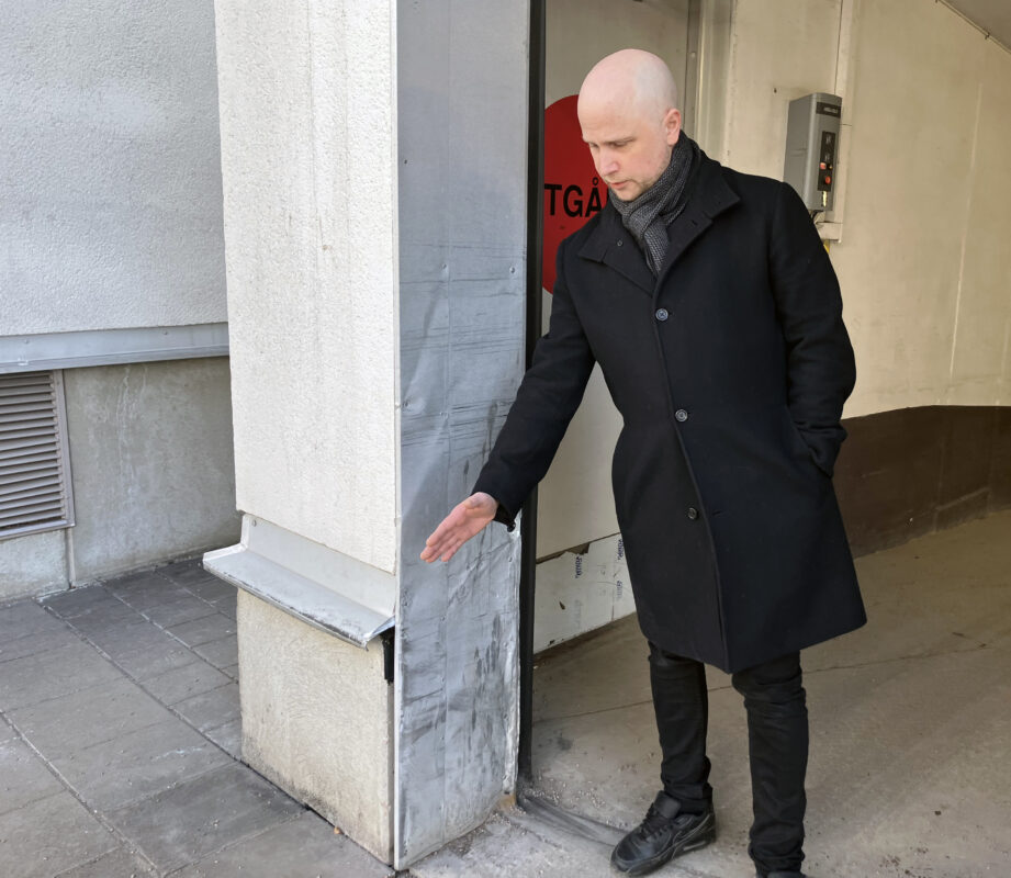 En man i svart rock och svarta byxor står i en garageöppning. Han pekar med ena handen mot ena pelaren. Han har väldigt lite hår och är barhuvad.