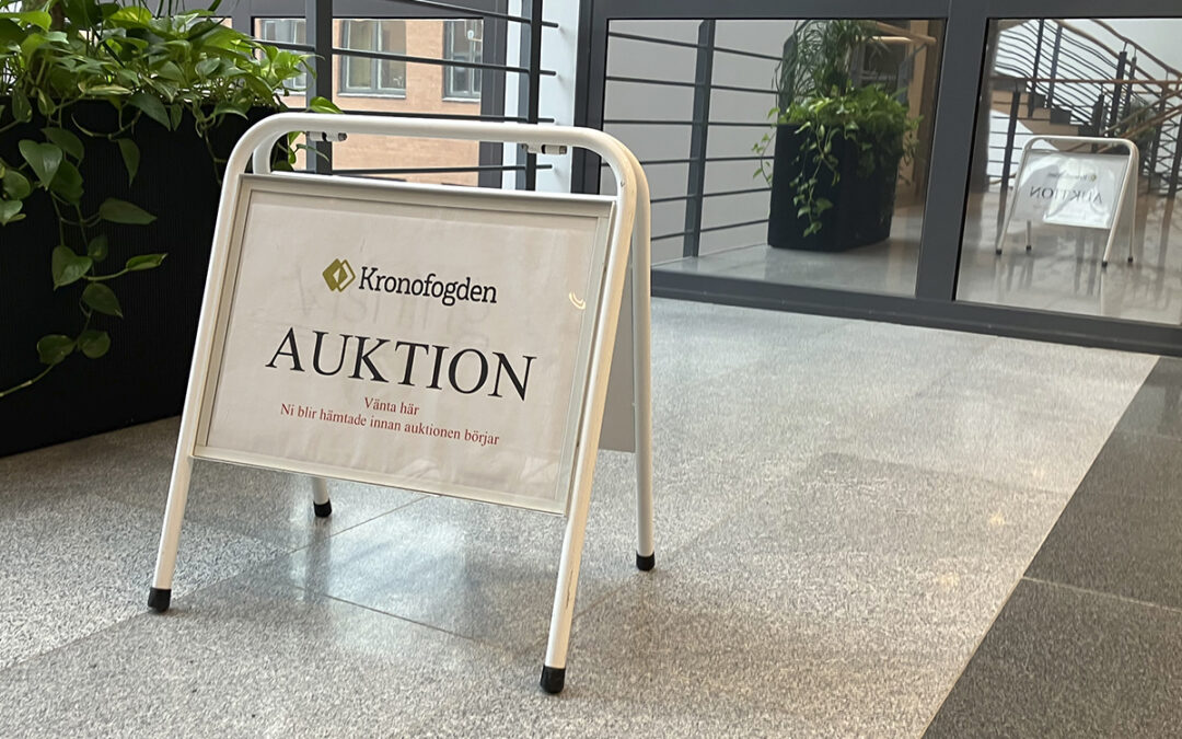 Skylt i gatupratarformat med Kronofogdens logotyp överst, texten "AUKTION" i versaler och under i röd text "Vänta här Ni blir hämtade när auktionen börjar". Till vänster gröna växter i en stor kruka, i bakgrunden spglas skylten och växterna i glasdörrar. Kronofogden försökte sälja några av AB-hems hyreshus i Sala, men buden som kom in var för låga för att någon försäljning skulle bli av.