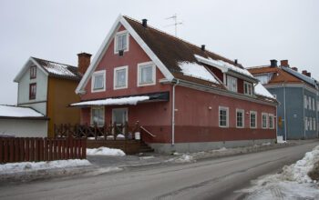 Ett ljusrött putsat hus med två våningar och en vindsvåning, på andra sidan en gata. Snö på marken och delar av taket. I bakgrunden ett ljusblått något högre hus. Kronofogden är på väg att tvångssälja några av AB-hems hus i Sala.