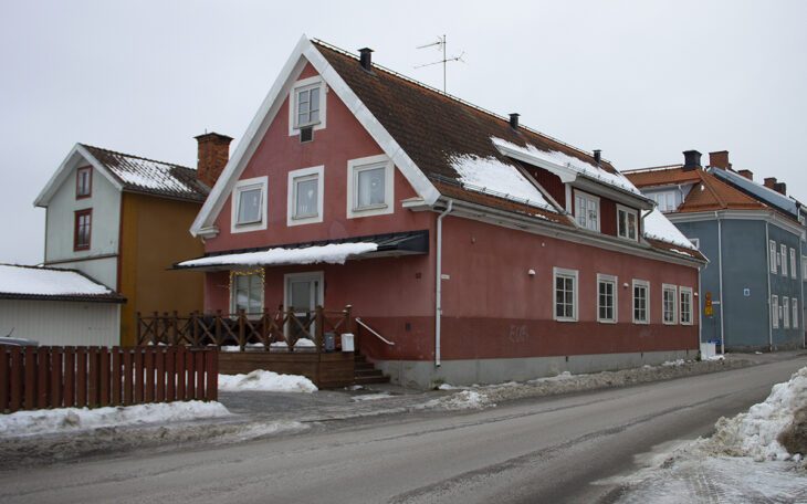 Ett ljusrött putsat hus med två våningar och en vindsvåning, på andra sidan en gata. Snö på marken och delar av taket. I bakgrunden ett ljusblått något högre hus. Kronofogden är på väg att tvångssälja några av AB-hems hus i Sala.