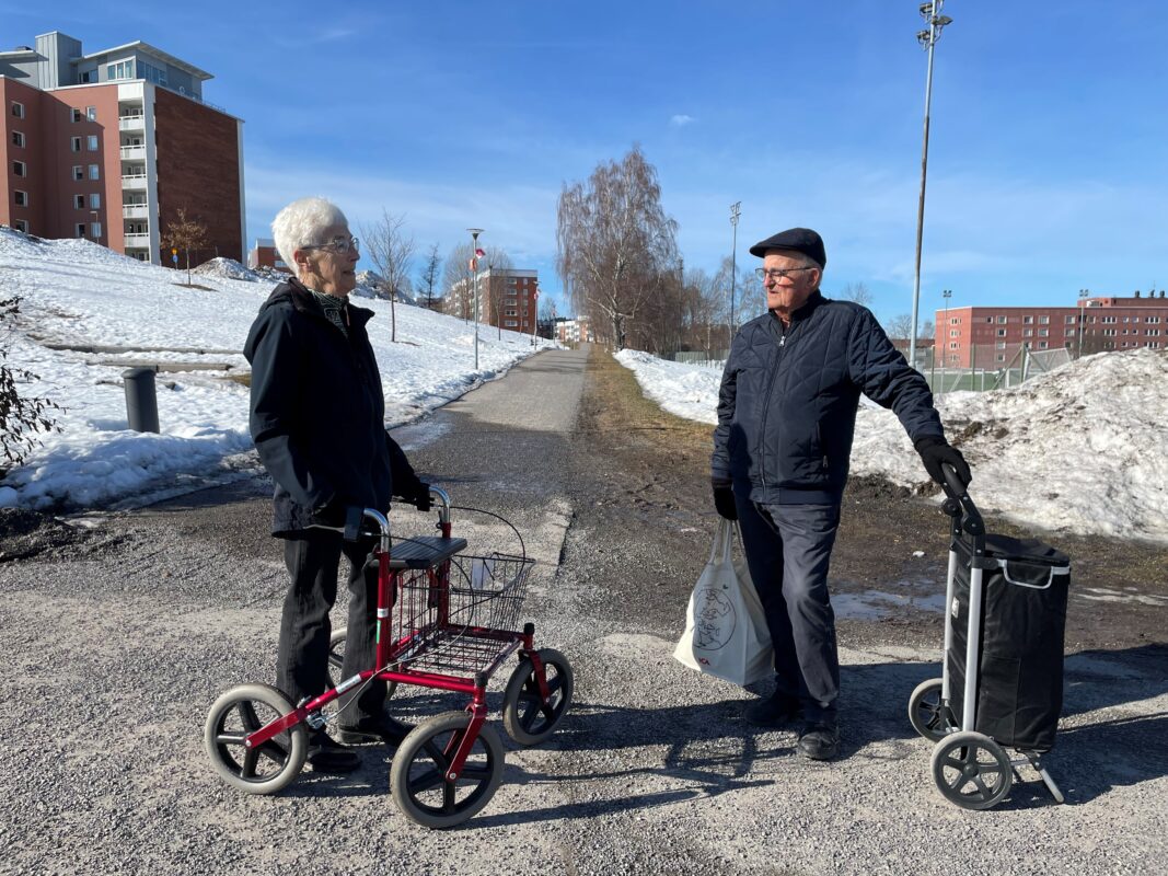 En äldre dam och en äldre herre står utomhus och samtalar på en gångväg i ett bostadsområde. Damen har en rollator och mannen en så kallad dramaten.