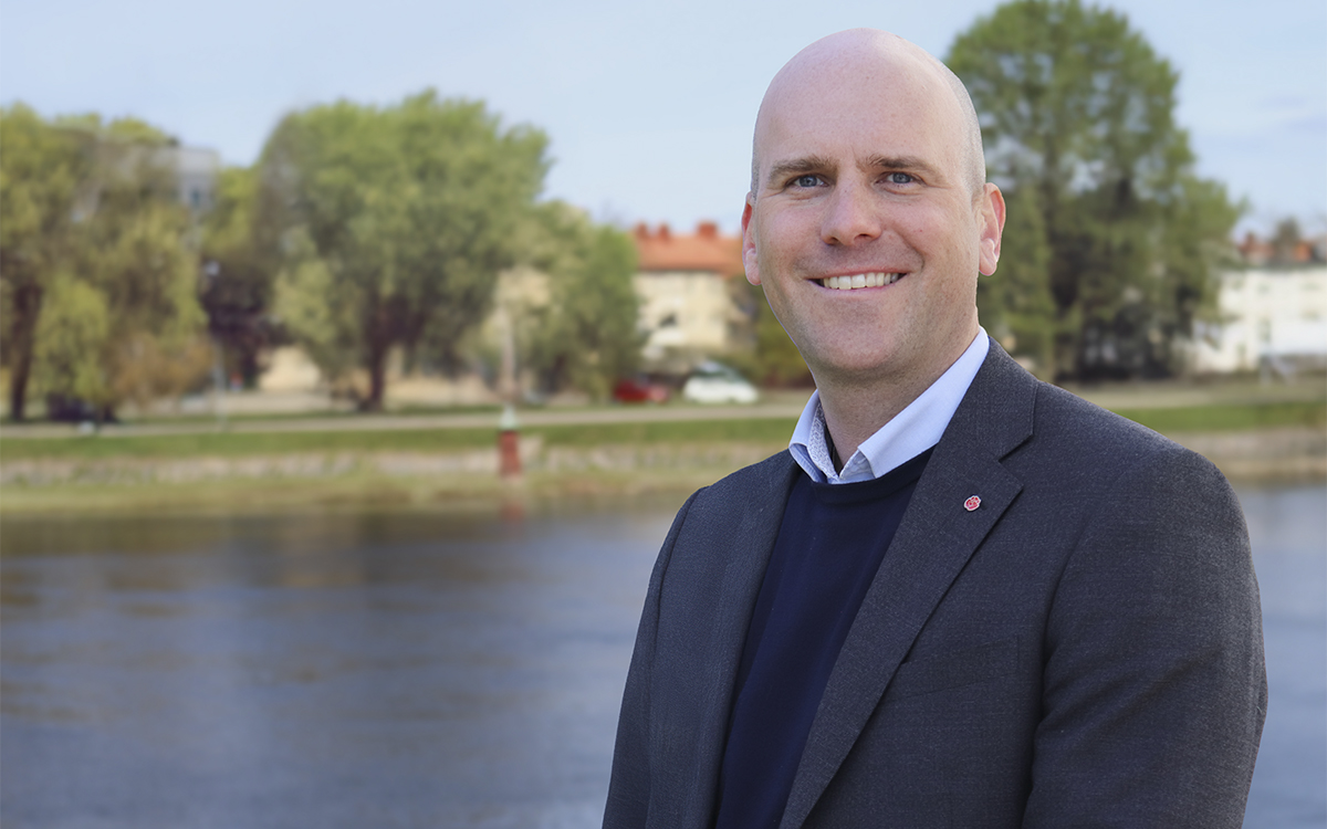Halkroppsporträtt på Anders Tallgren i mörk kavaj och pullover, ljus skjortkrage som sticker fram. Rakat huvud, liten röd ros - Socialdemokraterna-pin - på kavajslaget. I bakgrunden en älv och på andra sidan träd, gräsmatta och bsotadshus, Anders Tallgren (S) försvarar Karlstads kommuns värdeöverföringar från KBAB.