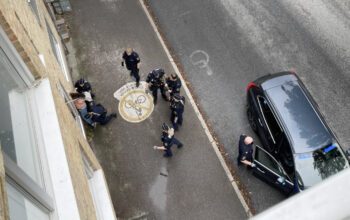 Bild tagen uppifrån från en balkong. Två poliser trycker upp en man med bar överkropp mot en vägg. På trottoaren syns ytterligare fyra poliser. Samt en civil polisbil som är parkerad på gatan.