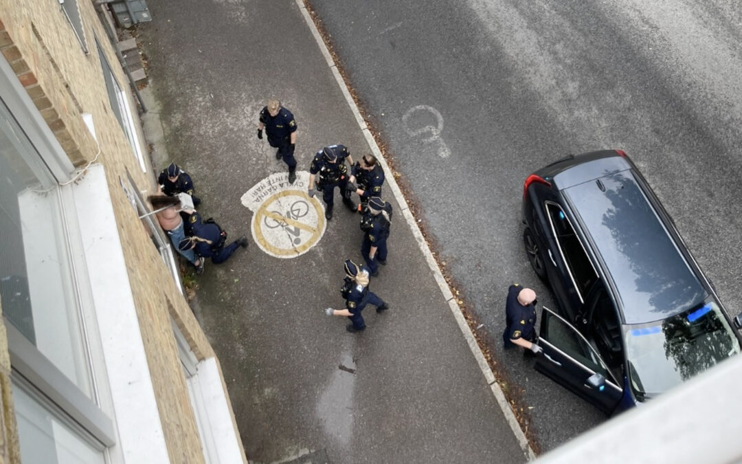 Bild tagen uppifrån. Poliser omringar en man med bar överkropp och trycker upp honom mot en vägg. På gatan en civil polisbil.