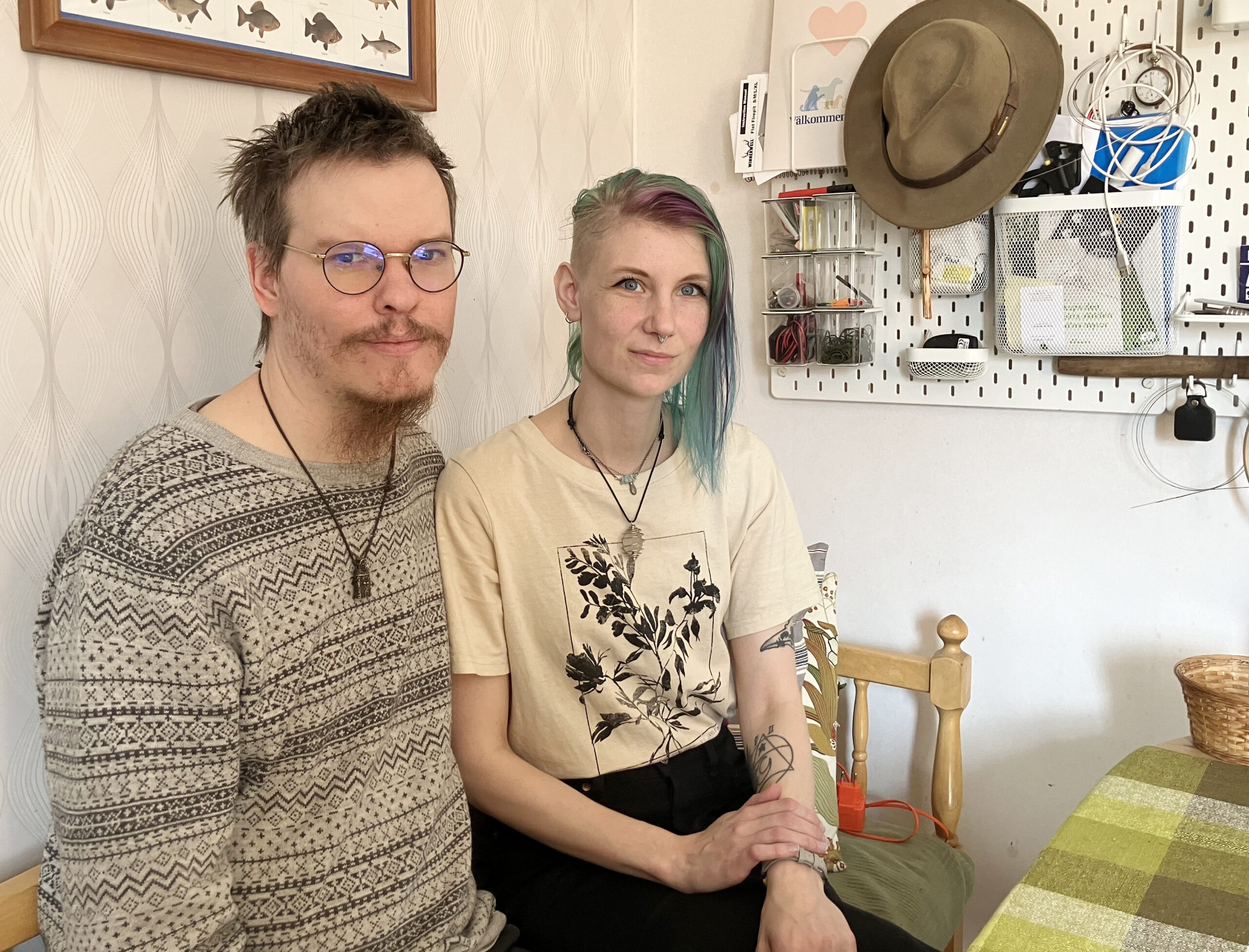 En man och en kvinna i 30-årsåldern sitter bredvid varandra på en soffa. Han har kort spretigt hår, glest skägg, mustasch och glasögon. Randig tröja. Hon har kort hår på ena sidan och längre på andra - färgat i lila och grönt. T-shirt med tryck.