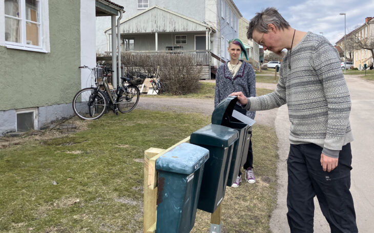 En man i 30-årsåldern står vid en rad med brevlådor och lyfter upp locket till en av dem. Han har kort hår, ett litet glest skägg och glasögon. Randig långärmad tröja och svarta byxor. I bakgrunden står en kvinna i samma ålder och tittar på honom. Hon har en gråmelerad kofta och hår som är kort på ena sidan. På andra sidan långt och går i grönt och lila. I bakgrunden ett hus med grönrappad fasad.