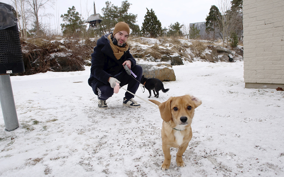 I förgrunden: En ganska liten hund med blond päls. Bakom sitter Dino Norberg på huk och håller i kopplet till den och ytterligare en hund, svart, i bakgrunden. Han har på sig ljusbrun mössa och halsduk, mörkblå vinterjacka och adidasbyxor, Dino Norberg är hyresgäst och bor i Hammarö kommun. Han är nöjd med att kommunen inte tar ut pengar från bostadsbolaget.