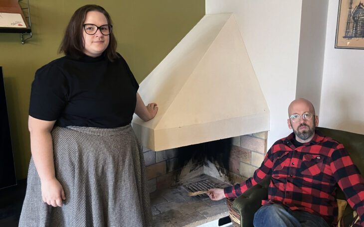 En kvinna står vid en öppen spis och en man sitter i en fotölj vid den. Kvinnan står t v i bilden och håller sin hand på spisen. Hon har mörkt axellångt hår, glasögon, svart kortärmad topp och vid grå kjol. Mannen har jeans, rödrutig skjorta och glasögon.l Är tunnhårig. Han håller i en vedklabb som han sträcker in i den tomma spisen, och tittar allvarligt in i kameran, liksom kvinnan.