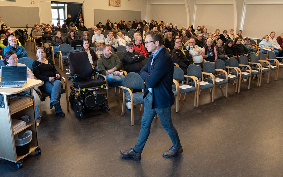 En man som går framför en samling människor som sitter ned på stolar i en samlingssal.