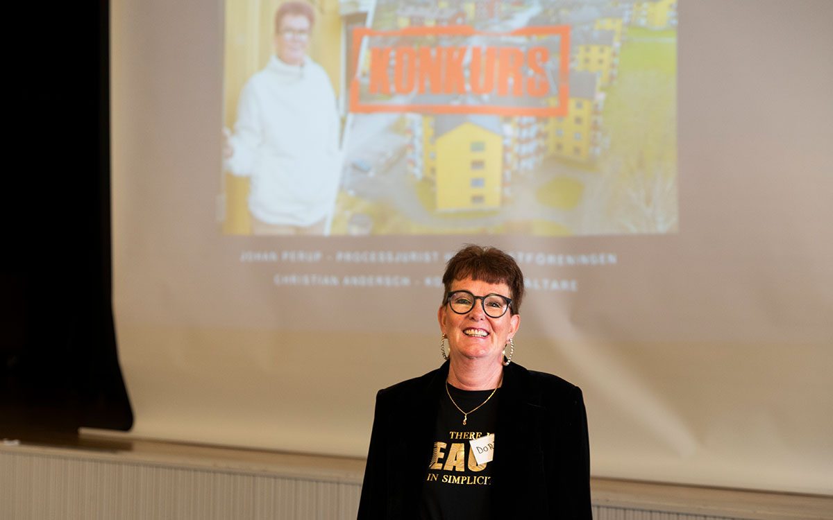 En leende kvinna i kavaj och en namnskylt med texten Doris som står framför en projicerad bild på henne själv, gula hus och en stämpel med texten konkurs.