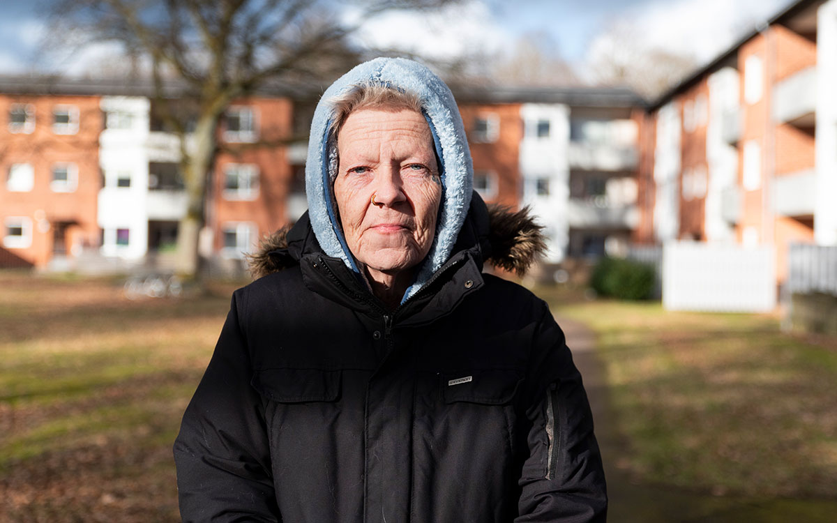 En äldre kvinna med blåa ögon, tatuering vid ena ögat, piercing i näsan och blå luva uppdragen över huvudet.
