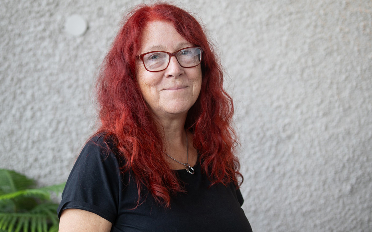 Halvkroppsbild på medelålders kvinna med långt tjockt eldrött hår, röda glasögon och svart t-shirt. Hon tittar in i kameran och ler lätt med stängd mun. 