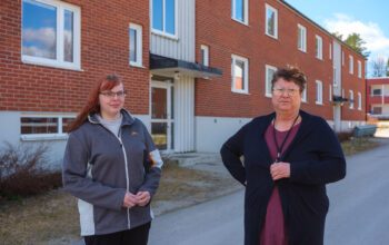 Två kvinnor framför ett flerfamiljshus i rött tegel med två våningar. Kvinnan t v har rött långt hår, glasögon, en grå träningsoverallsjacka och håller händerna knäppta framför sig. Den andra har kort mörkt hår, lång svart kofta, lila klänning. De tittar båda in i kameran.