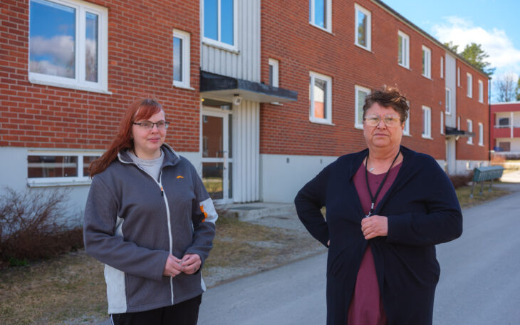 Två kvinnor framför ett flerfamiljshus i rött tegel med två våningar. Kvinnan t v har rött långt hår, glasögon, en grå träningsoverallsjacka och håller händerna knäppta framför sig. Den andra har kort mörkt hår, lång svart kofta, lila klänning. De tittar båda in i kameran.