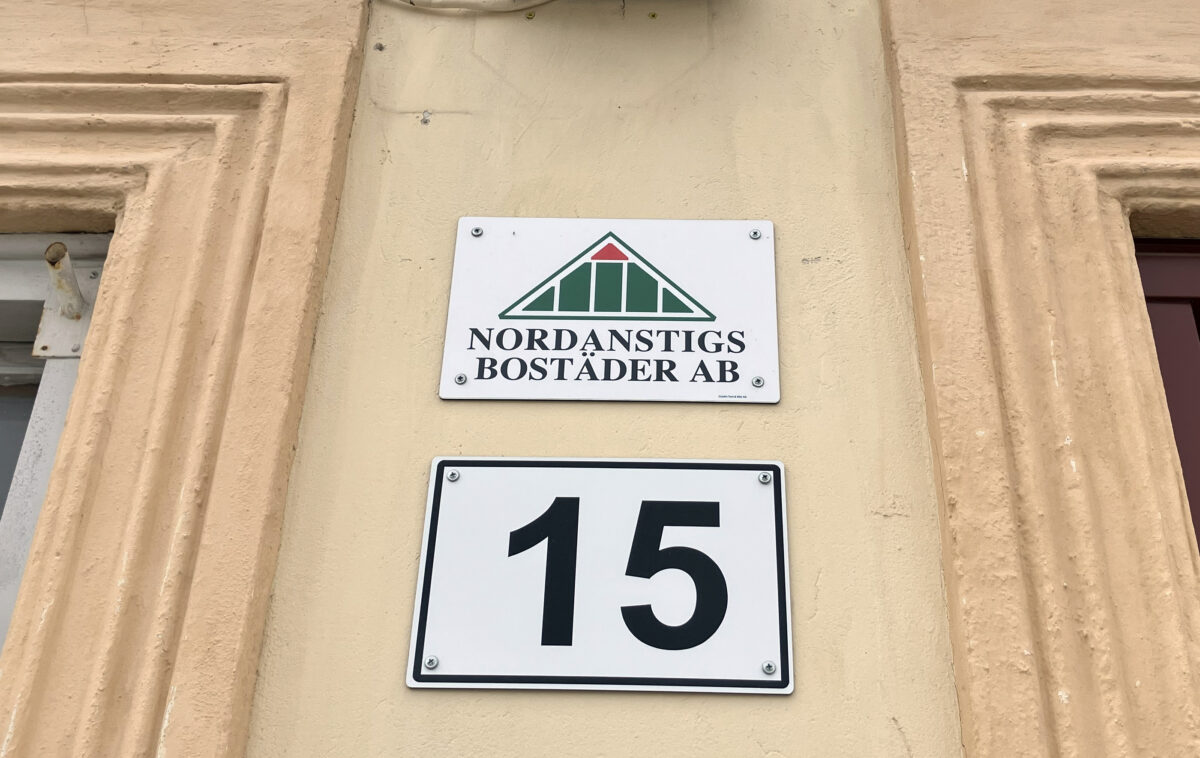 Nordanstigs bostäders skylt mot en svagt gul vägg. Under den en skylt med siffran 15.