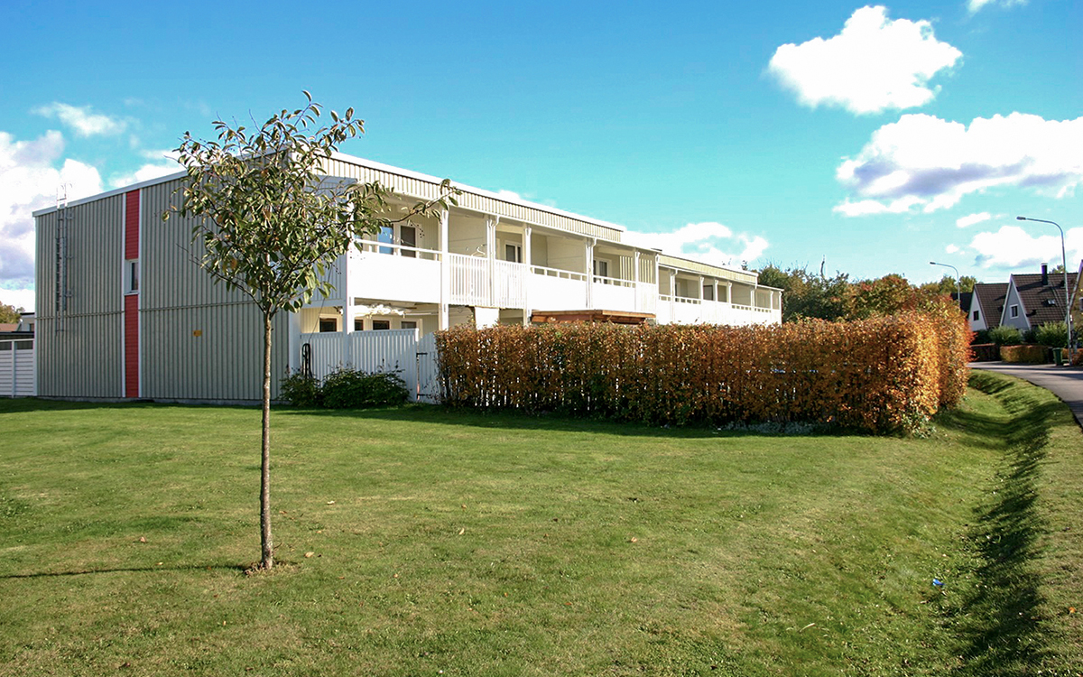 Bilden visar en vit radhuslänga. I förgrunden en gräsmatta, därefter syns häckar som ser ut att omgärda uteplatserna som ses framför radhuslägenheterna.