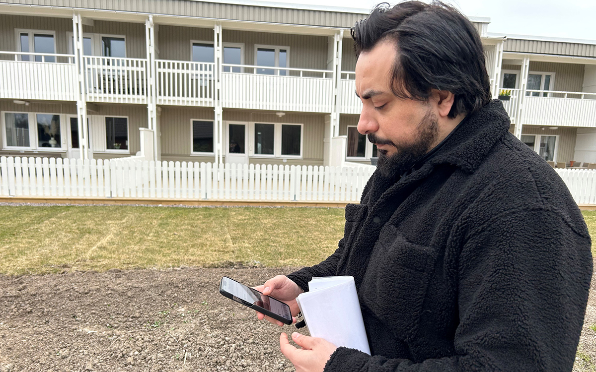 Man i profil med en mobiltelefon i sin hand. Han tittar ner på den. I bakgrunden syns en remsa med jord och därefter följer en gräsremsa. Bakom det ett vitt radhus i två våningar.