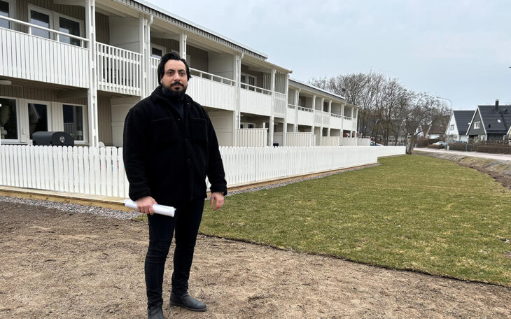 Man som står framför ett radhus. Han håller en rulle med A 4-ark i ena handen. Ett lågt, vitt staket syns bakom honom samt en gräsmatta. Staketet skärmar av tomterna och gräsmattan går ut mot vägen .