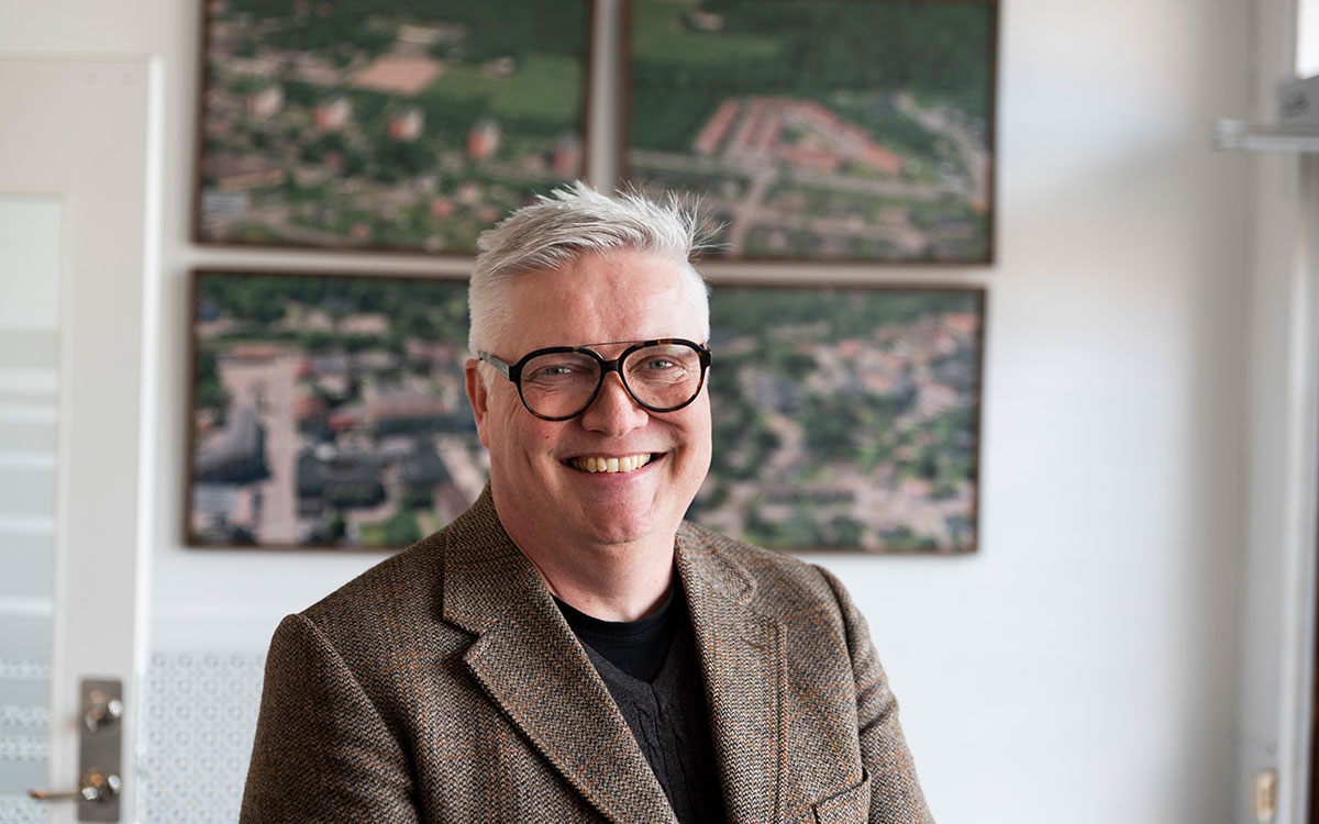 En man med glasögon, vitt hår och kavaj i ett kontor framför flygbilder på bostadsområden.