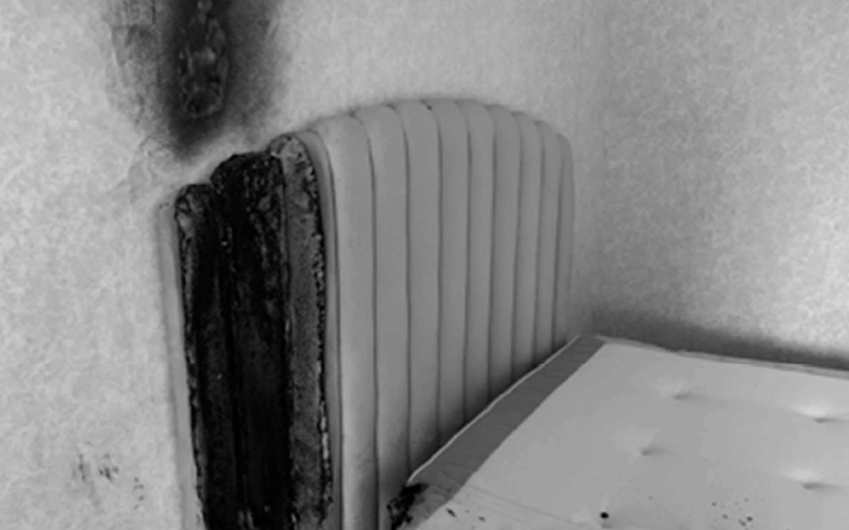 Svartvit bild som visar att det har brunnit i kanten av sänggaveln. Dessutom ser man en del av tapeten där det har brunnit.