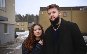 Halvkroppsporträtt i miljö. Shandi Moen till vänster i brund vågigt hår och svart täckjacka, står tätt intill Matti Klenz-Tornow i lite lkusare kortklippt frisyr, mörkt helskägg och liten mustasch, svart jacka, tjock guldkedja om halsen, ett litet örhänge i vänster öra. I bakgrunden flerfamiljshus i gulbeige tegel med bruna tak. Snö på marken. Shandi Och Matti tycker att det är konstigt och orättvist att en del av deras hyrespengar går till kommunen.