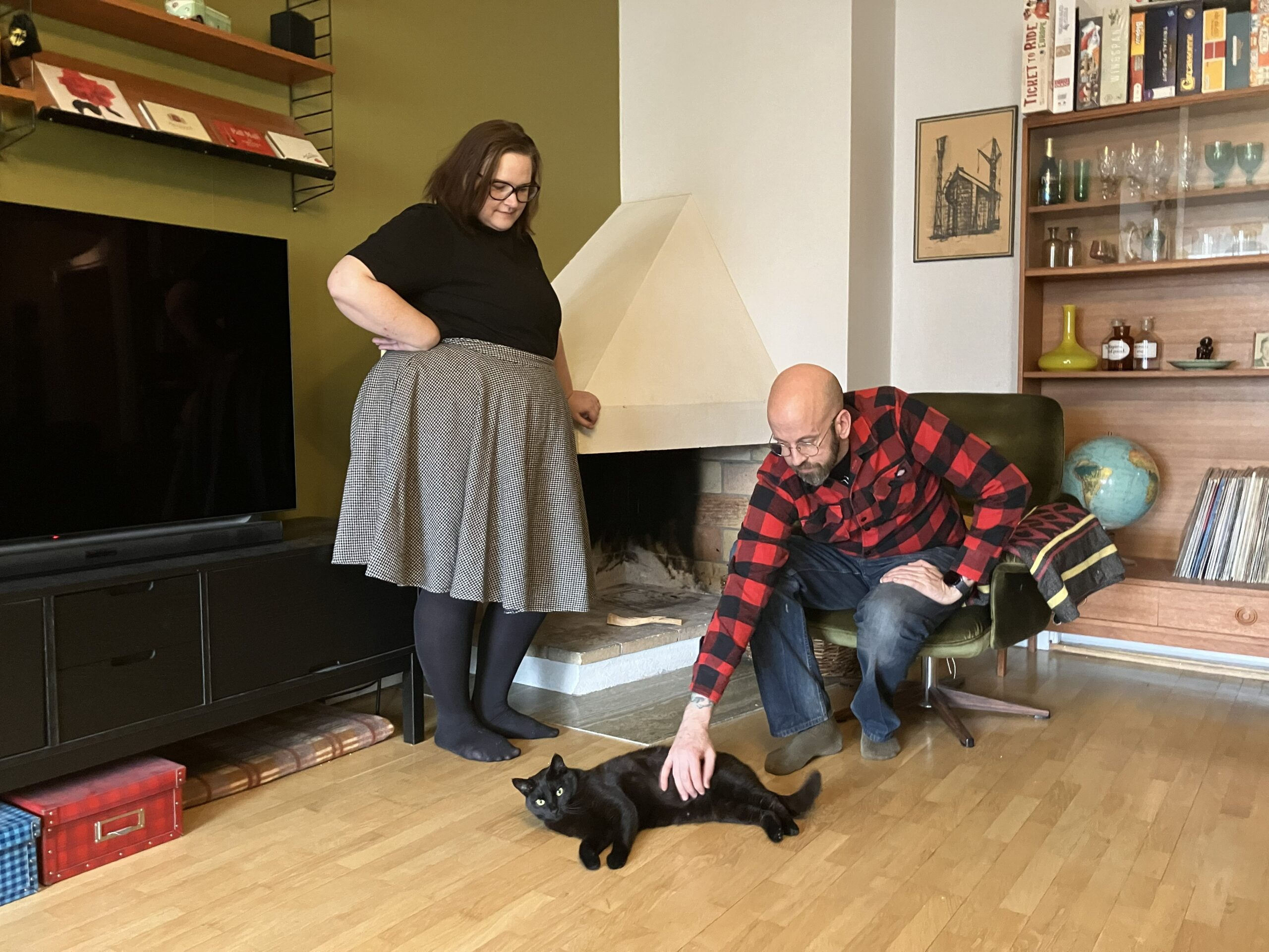 Man, kvinna och katt vid öppen spis. Kvinnan håller handen på spiskåpan och tittar ner på katten på golvet. Hon har mörkt axellångt hår, svaqrt topp och grå vid kjol. Mannen är kal på huvudet, har skägg, glasögon, rödsvartrutig skjorta och jeans. Han klappar den svatrta katten.