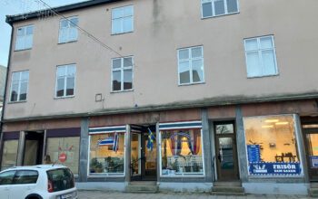Bilden visar ett flerfamiljshus i tre våningar. I bottenvåningen är det affärslokaler. Framför huset står en vit bil.