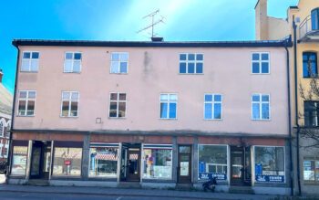 Bilden visar ett flerfamiljshus i tre våningar. I bottenvåningen är det affärslokaler.