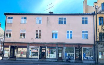 Bilden visar ett flerfamiljshus i tre våningar. I bottenvåningen är det affärslokaler.