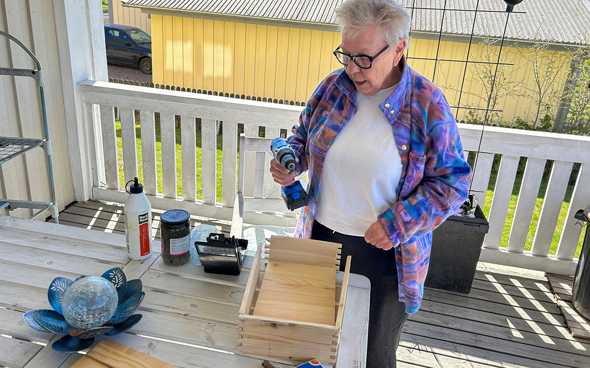 En kvinna som håller en borr i sin hand. På ett bord framför henne ligger en trälåda som inte är färdig – sidorna är inte sammanfogade. På bordet syns även en ljuslykta och en flaska med trälim.