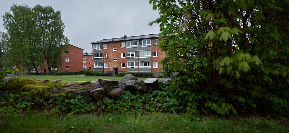 Rött tegel med trevånings flerbostadshus bakom en stenmurad trädgård och grönska.