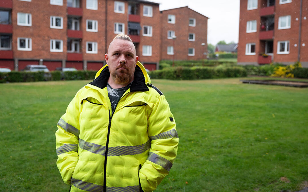 En man med mohaikanfrisyr, skägg och en gul varseljacka som står framför röda tegelhus.