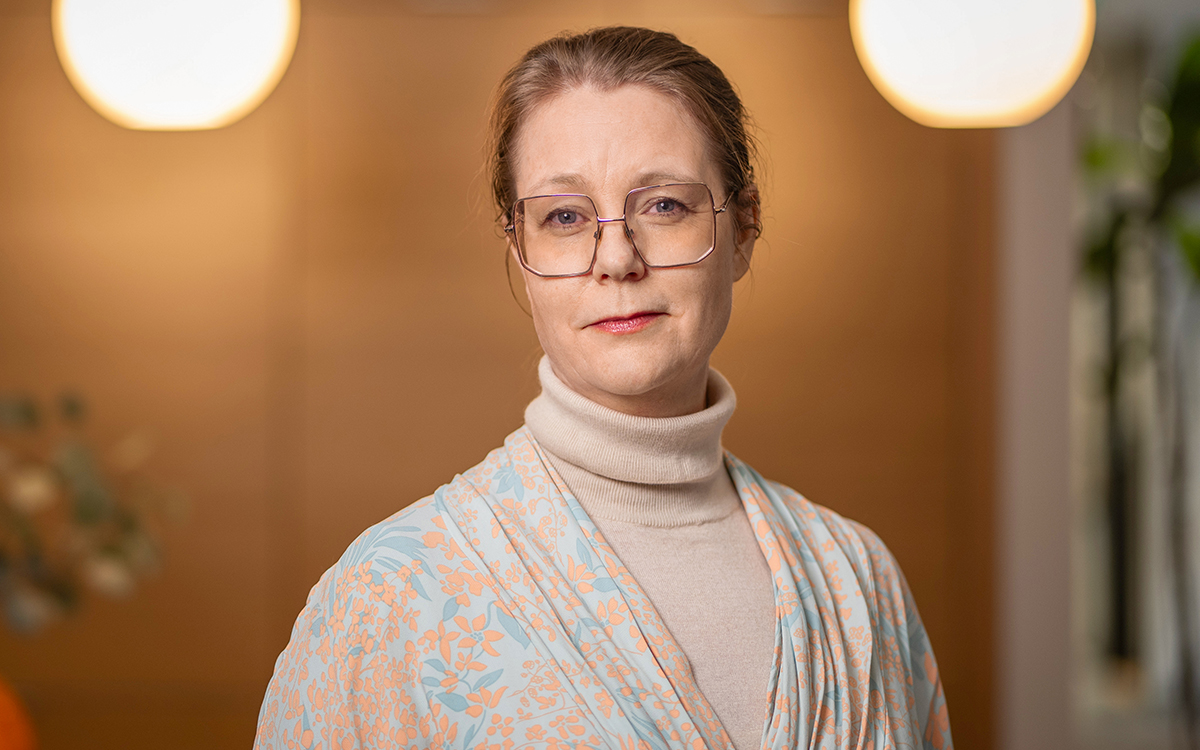 Bild är tagen rätt framifrån på en kvinna som tittar in i kameran. Halvfigur. Två klotformade lampor syns lite svagt i bakgrunden.