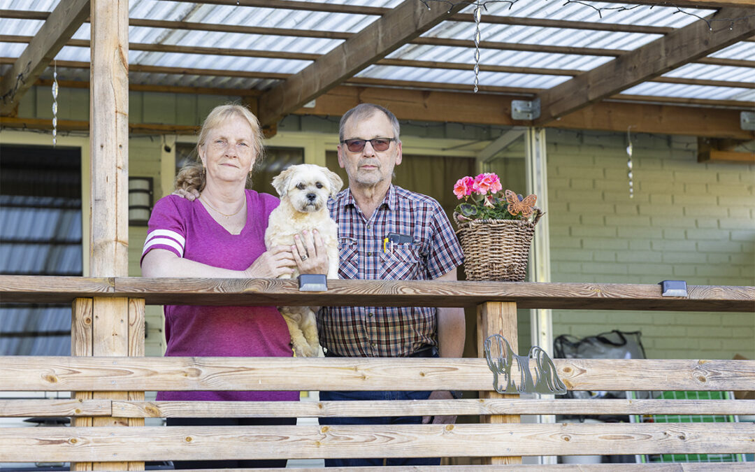 Eva (i rosa kortärmad tröja och långt ljust hår i hästsvans) och Bonny Isaksson (i rutig kortärmad skjorta, kort grått hår och solglasögon) står bakom ett altanräcke, på räcket mellan dem håller Eva upp den lilla hunden Bella som har ljus päls. Till höger om dem en stor korg med en rosa pelargon. Ovanför dem ett altantak som släpper igenom lite ljus. Eva och Bonny Isaksson upptäckte att de betalat el och vatten åt grannen där de bodde tidigare.