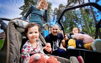 En småbarnsfamilj. Närmast kameran en liten flicka som ler och tittar in i kameran. Stående bakom henne mamman. Sittande på huk gör pappan. Han håller i handtaget till en kärra där ytterligare en liten flicka ligger.