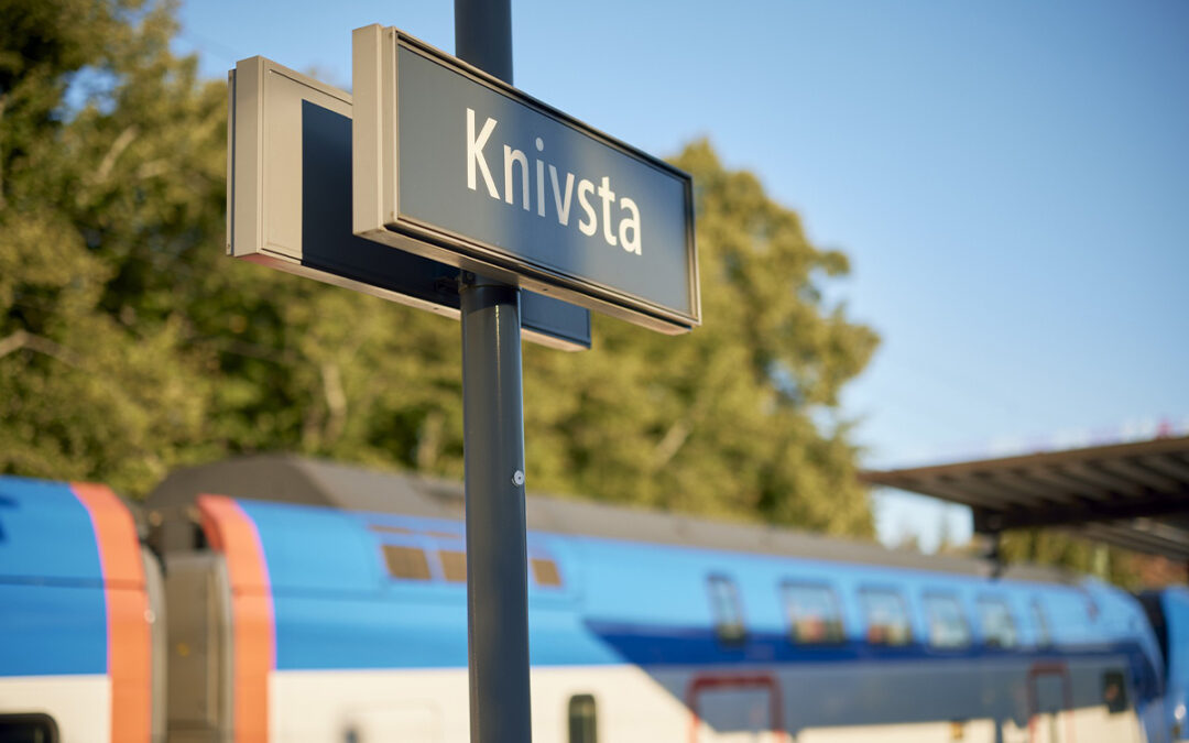 Bilden föreställer en järnvägsskylt som det står Knivsta på. I bakgrunden ett SL-tåg, bakom det grönska.