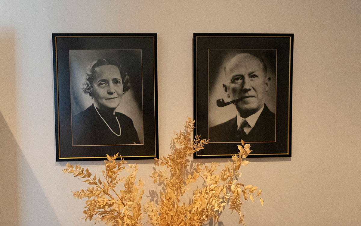 Två svartvita fotografier på en kvinna med pärlhalsband samt en man med pipa på en vägg bakom en torkad blomma.