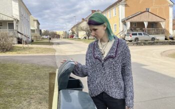 Halvkroppsbild på Carolina Södergran, med axellångt hår till stor del färgat i grönturkos. Hon har på sig en stickad tröja och tittar ner i en brevlåda som hon har öppnat- I bakgrunden en gata med flerfamiljshus i två våningar som ser ut att kunna vara runt 100 år gamla. Carolina bor hos AB-hem i Hofors. I våras slutade Postnord dela ut deras post tillfälligt för att AB-hem inte hörsammat ett krav på att flytta brevlådorna. Förra veckan hotade fjärrvärmabolaget med att stänga av värme och varmvatten, för att AB-hem inte betalat räkningarna i tid.
