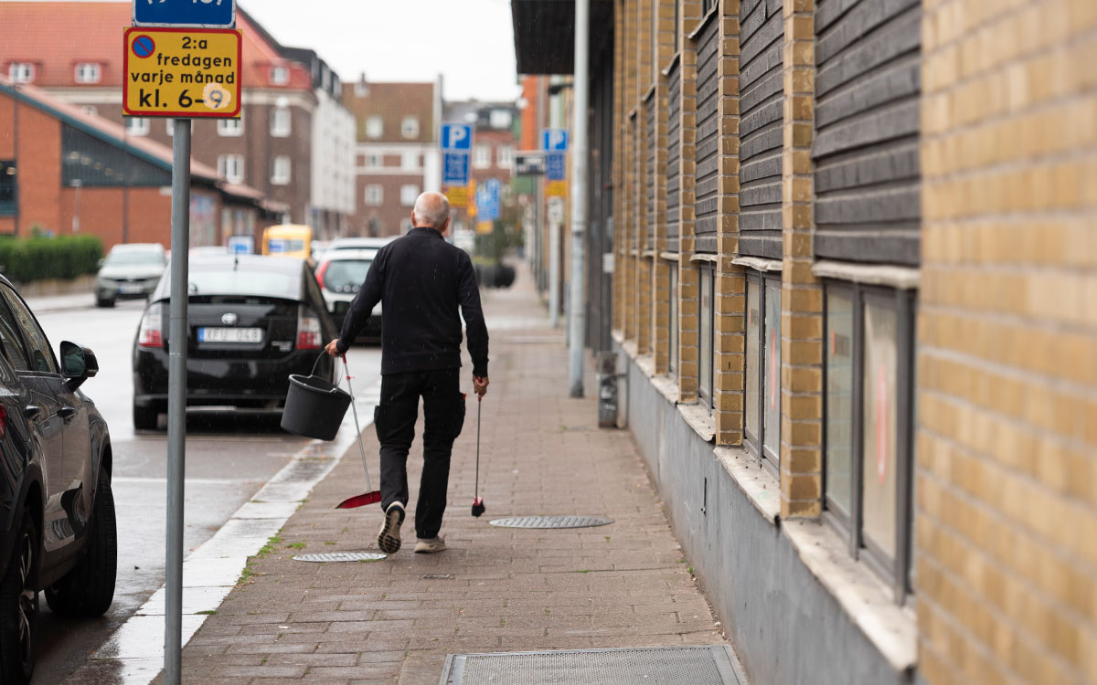 En äldre man med vitt hår och arbetshandskar som går och sopar utanför ett gult tegelhus.