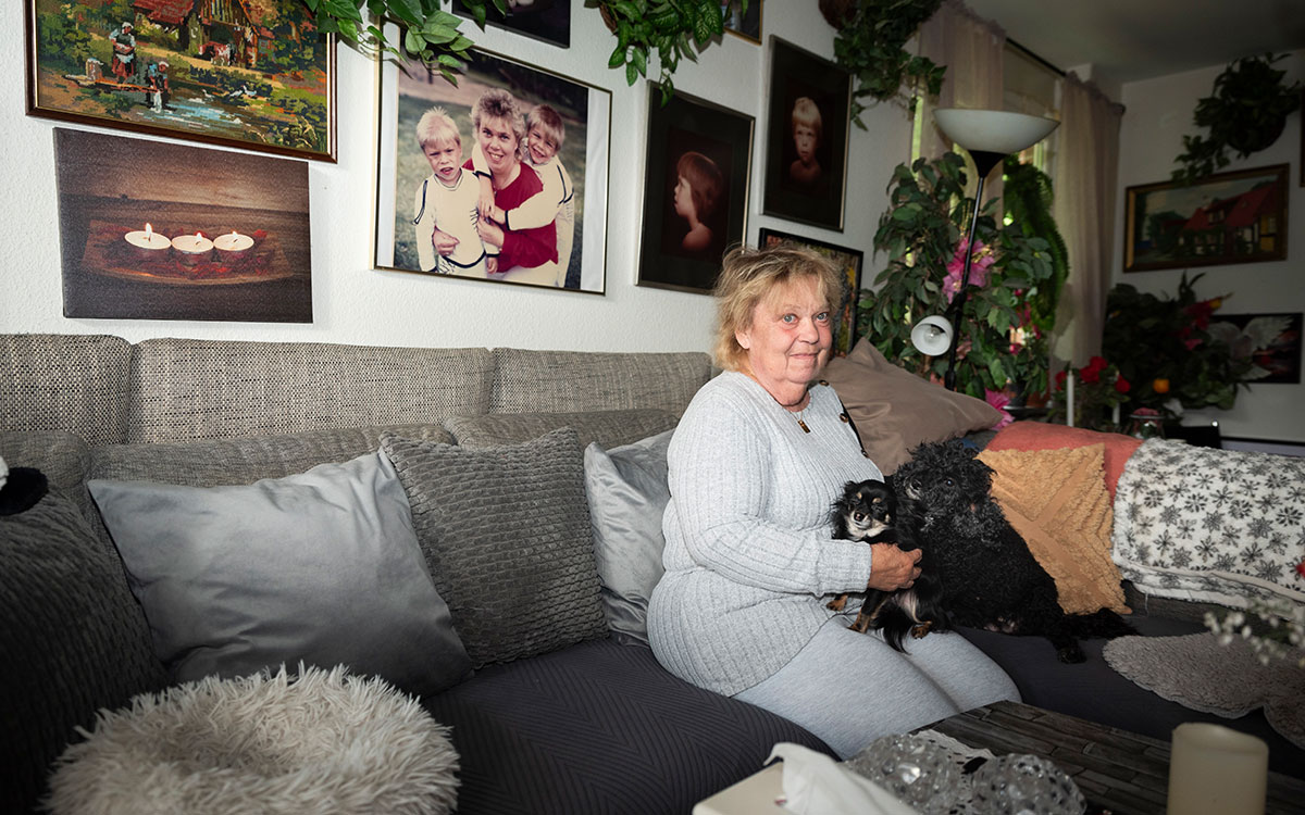 En äldre kvinna som sitter i en soffa med en liten hund i ett vardagsrum och på väggen bakom finns fotografier på släktingar.