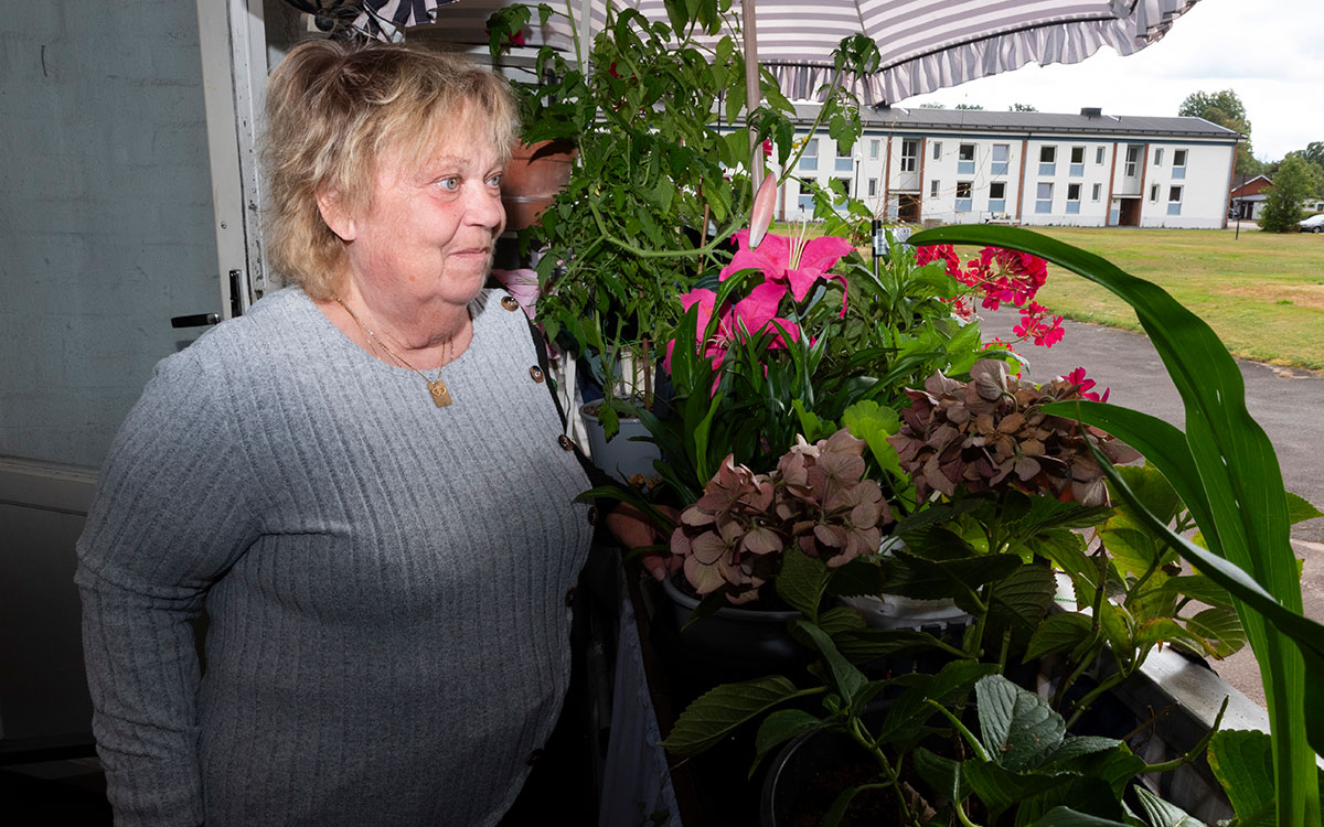 En äldre kvinna som står på en veranda med många växter och ett flervåningshus och en gräsplan i bakgrunden.