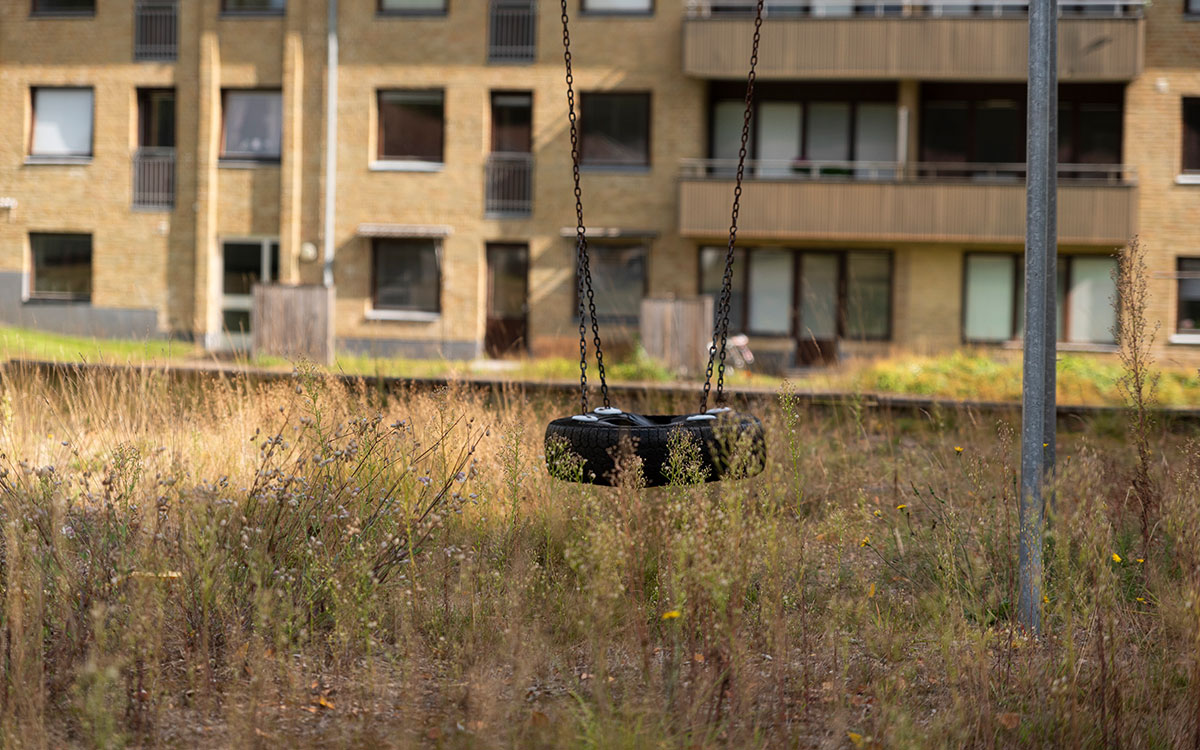 En gungställning och en helt igenväxt sandlåda framför ett flervåningshus i gult tegel.