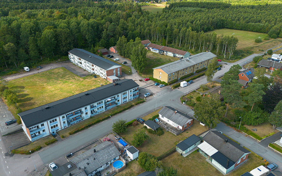 En bild tagen ovanifrån som visar tre större huskroppar samt marklägenheter i ett bostadsområde om givet av skog och villabebyggelse.