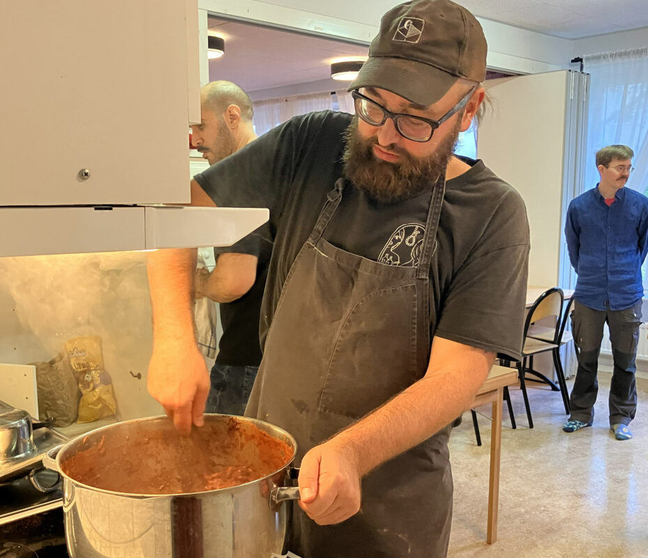 En man med mörkt hår och skägg och mörka glasögon står vid en spis och rör i en chiligryta. Han har keps, svart t-shirt och förkläde. I bakgrunden syns två män.